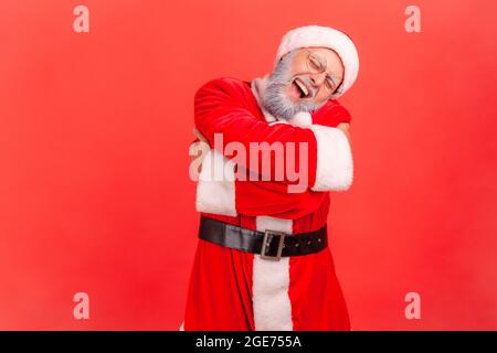 Elderly man with gray beard wearing santa claus costume hugging himself, feeling comfortable, narcissistic egoistic person, happy expression. Indoor s Stock Photo