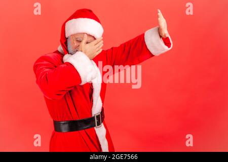 Side view portrait of elderly man with gray beard wearing santa claus costume standing, covering his face and showing stop hand gesture. Indoor studio Stock Photo