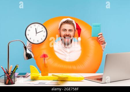 Happy excited man in santa claus hat sitting at workplace with toy plane in rubber ring on neck, showing passport and wall clock, time to go on winter Stock Photo