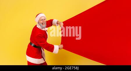Side view portrait of elderly man with gray beard wearing santa claus costume carrying big bag with presents, copy space for advertisement. Indoor stu Stock Photo