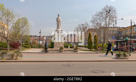 Mladenovac, Serbia - April 13, 2020: People With Face Masks Walking by WWI Soldier Monument Landmark in Park. Stock Photo