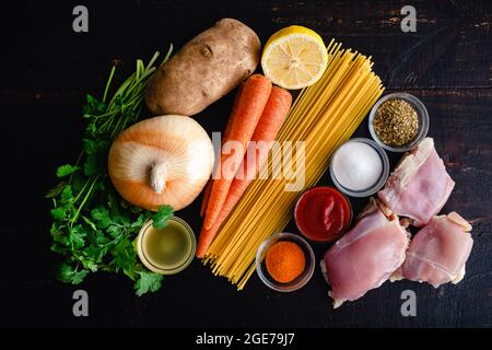 Ingredients for Cuban Style Chicken Noodle Soup on a Dark Wood Background: Chicken thighs, spaghetti noodles, vegetables, and spices on a wooden table Stock Photo