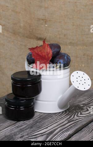 Large plums in a metal watering can. On pine boards. Nearby is a jar of jam. Harvest plums. Stock Photo