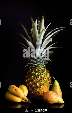 Still life of fruit in which there is a pineapple with its leaves, some pears and some bananas, on a dark background. The fruit helps to keep you heal Stock Photo