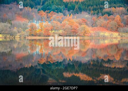 A calm, peaceful lake reflection of vibrant, autumn colours and a waterfront stately home on Loch Achray in Loch Lomond and the Trossachs National Par Stock Photo