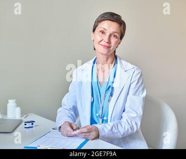 Smiling middle-aged woman doctor with stethoscope on shoulders in white coat working in clinic Stock Photo