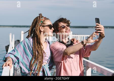 Smiling stylish friends in sunglasses standing on pier and taking selfie on smartphone Stock Photo