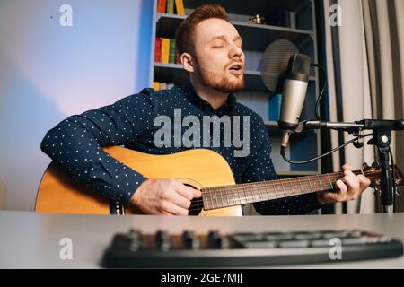Close-up shoot from below of guitarist singer man play on acoustic guitar recording sing song using professional microphone stand at home studio. Stock Photo