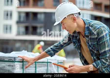 Serious concentrated worker in a hardhat checking construction materials outdoors Stock Photo