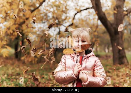 Little blonde girl plays with yellow autumn leaves in the garden, smile, has fun Stock Photo
