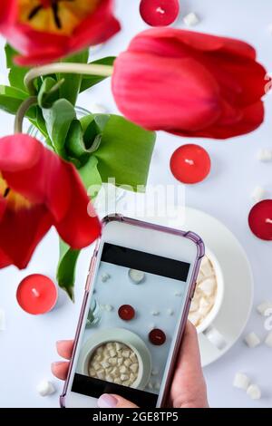 Red tulips candles White cup of cacao marshmallows morning breakfast. Female hand taking photo. Blogger. Valentines Womans Mothers day. Top view. Flat Stock Photo