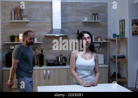 Abused woman sitting in kitchen with drunk husband dealing with aggression, conflict and alcohol addiction. Hurt bruised person having marriage problems with domestic violence Stock Photo