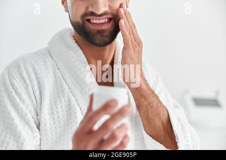 Cheerful bearded man in bathrobe applies moisturizing cream onto face on light background Stock Photo