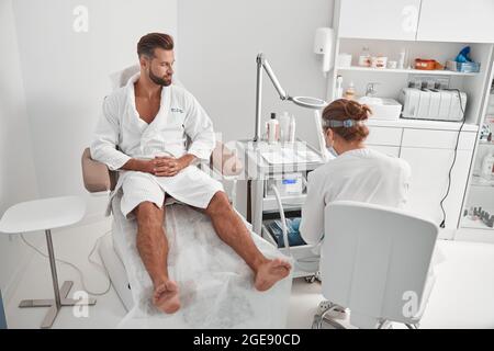 Man client in bathrobe sits in armchiar while master prepares instrument for pedicure procedure in salon Stock Photo