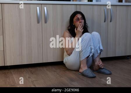 Close up of crying wife being victim of physical trauma and domestic violence. Miserable woman with bruises on face sitting on floor unhappy with aggressive husband and marriage Stock Photo