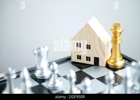 Wooden house model with a gold king chess on a chessboard with silver chess pieces surrounding it. Stock Photo