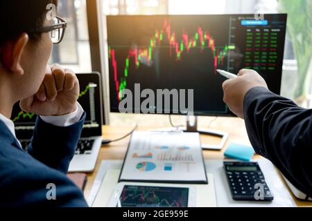 Businessmen are pointing to monitor to view stock market data on desktop computer and discuss it by analysis data from data graph. Stock Photo
