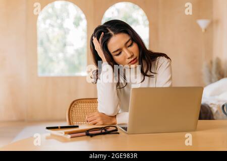 Exhausted ethnic female worker touching head and focusing on laptop screen while suffering from migraine during work at home Stock Photo