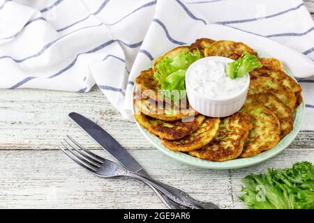 Fritters from zucchini with sour cream sauce and greens. Vegetable pancakes of courgettes with green salad on white wooden background. Stock Photo