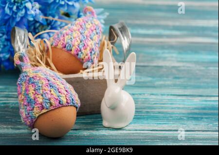 Easter composition with eggs in knitted hats, blue flowers and decorative rabbit on a wooden table. Stock Photo