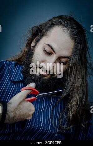 portrait of a young man with a beard and long hair who, with a scissor in his hand, hesitates to cut his hair, concept for willing change Stock Photo