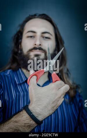 portrait of a young man with a beard and long hair who holds a scissor in his hand and looks at her while he thinks of cutting everything to change Stock Photo