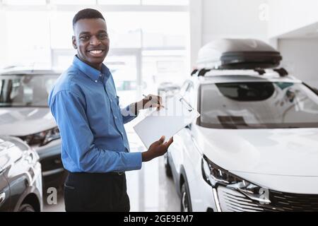 Cheerful black car salesman holding empty clipboard, smiling at camera in modern auto dealership, mockup Stock Photo