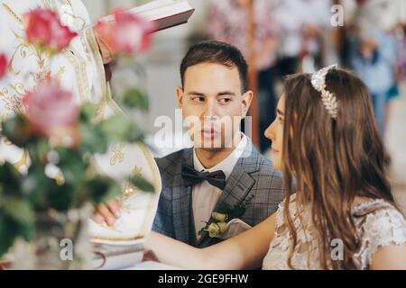 Stylish emotional bride and groom making oaths and holding hands on holy bible during matrimony in church. Wedding ceremony in cathedral. Classic wedd Stock Photo