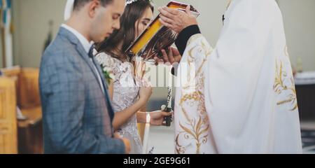 Stylish bride and groom kissing holy bible from priest hands during matrimony in church. Wedding ceremony in cathedral. Classic spiritual wedding coup Stock Photo