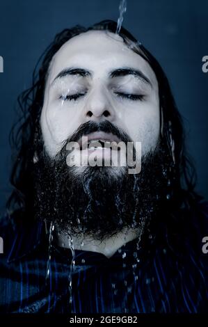 Portrait of a young bearded man who refreshes himself by pouring water on his head and running it on his face close up, blue toned image Stock Photo