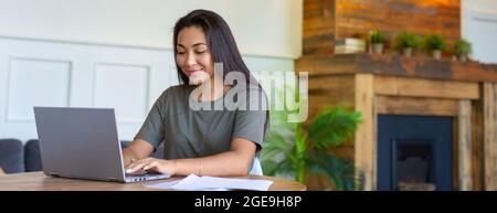 A woman works with papers at home in front of a laptop monitor. She is sitting with glasses and checking papers in fromt of laptop. Format 26x10. Stock Photo