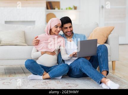 Loving pregnant arab spouses using laptop, sitting on floor at living room, watching baby room interior, free space Stock Photo