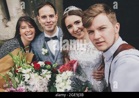 Stylish happy bride and groom taking selfie with family and smiling on background of church after holy matrimony. Beautiful emotional wedding couple w Stock Photo
