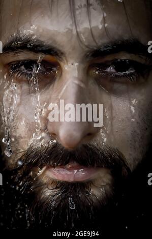 Very close-up portrait of a young bearded man with an intense gaze with streams of water running down his face Stock Photo
