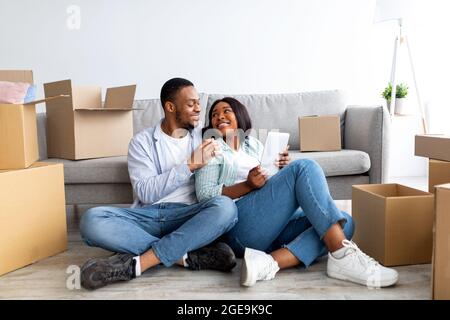 Loving black family using digital tablet and embracing while sitting on floor in new house among unpacked boxes Stock Photo