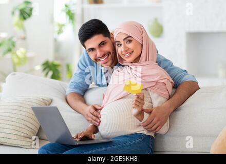 Preparing for childbirth. Happy muslim expecting family using laptop and credit card, shopping online at home Stock Photo