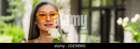 mature woman in yellow sunglasses holding white rose outdoors, banner Stock Photo