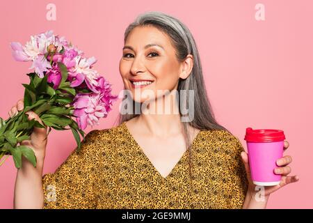 cheerful woman with flowers and coffee to go looking at camera isolated on pink Stock Photo