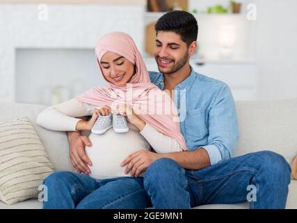 Happy muslim pregnant couple placing baby shoes on woman big belly while resting together on sofa at home Stock Photo