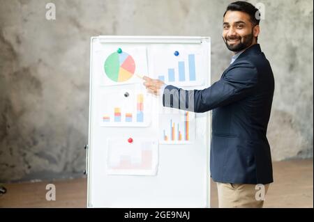 Cheerful Indian male tutor teaching students online, mixed-race man points at the whiteboard and looks at the camera, explaining, mentor holding webinar Stock Photo