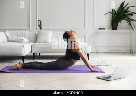 Concentrated young mindful indian woman in sportswear practicing yoga and watch online tutorial on laptop at home. Mixed-race female doing workout on mat with online class, listening tranquil music Stock Photo