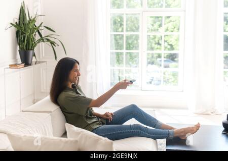 Side full length view of smiling indian woman lying on the comfortable white sofa with remote controller, watching TV, switching channels, spends lazy weekend in cozy modern living room Stock Photo