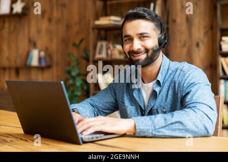 Smiling Indian male freelancer wearing headset and smart casual wear using laptop for connection with colleagues or customers, talking online sitting in the home office and looks at the camera Stock Photo