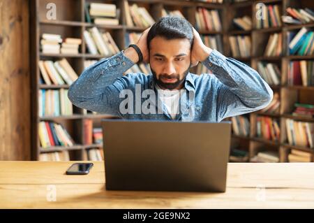 Amazed Indian male freelancer looks at the laptop monitor and holding head in despair, reading shock news, received bad notification sitting at the desk in the office Stock Photo
