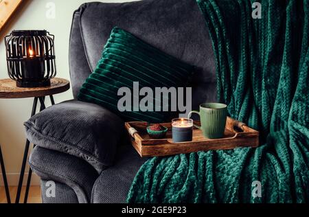 Modern autumn hygge set in living room. Dark green interior elements, soft pillows, plaid on sofa with chocolate muffin, aroma drink mug on wood tray Stock Photo