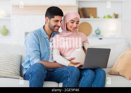 Loving pregnant muslim couple using laptop computer, resting on sofa in living room, spending time together, empty space Stock Photo