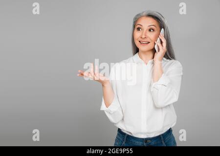 excited middle aged woman in white shirt gesturing while talking on smartphone isolated on grey Stock Photo
