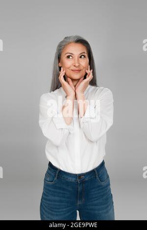 sly woman in white shirt touching face while looking aside isolated on grey Stock Photo