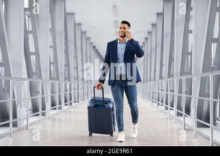 Handsome Arab Man Walking With Suitcase In Airport And Talking On Cellphone Stock Photo