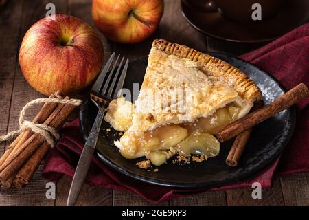 Slice of apple pie on a plate with apples and cinnamon sticks in background Stock Photo